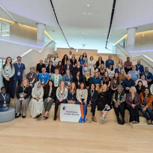 Alt text: A large group of diverse individuals posing on a wide indoor staircase in a modern building. The group includes mostly women, with a few men, all wearing name badges. A sign in front reads "Ford Women in Cyber Security."